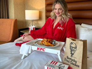 beautiful woman is in a kfc robe sitting on a hotel bed about to eat chicken and waffles